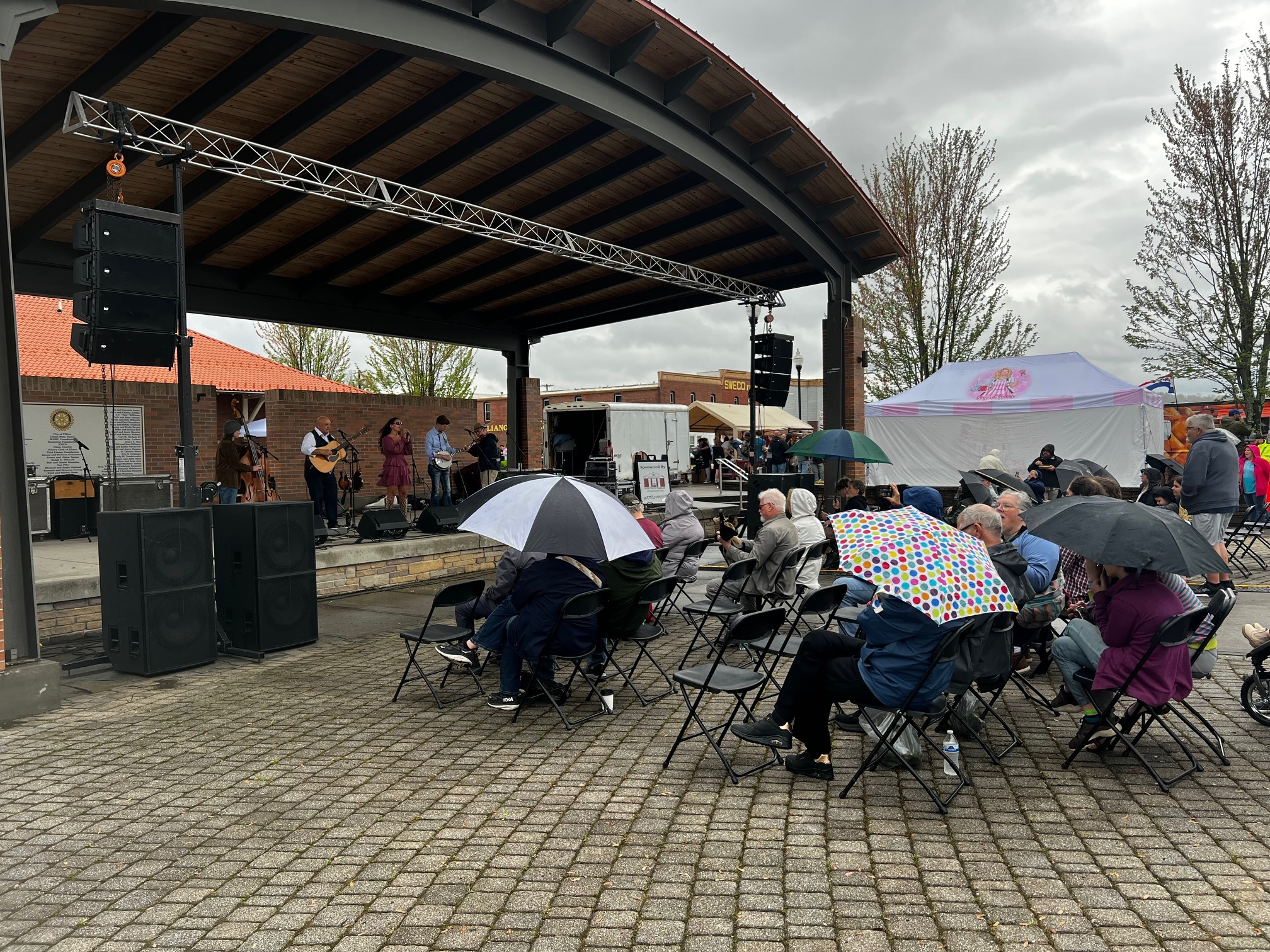 People sit on folding chairs under umbrellas, watching a band perform on an outdoor stage. The sky is cloudy and the ground is wet, indicating rain. Some attendees gather under a tent, while trees and buildings are visible in the background.