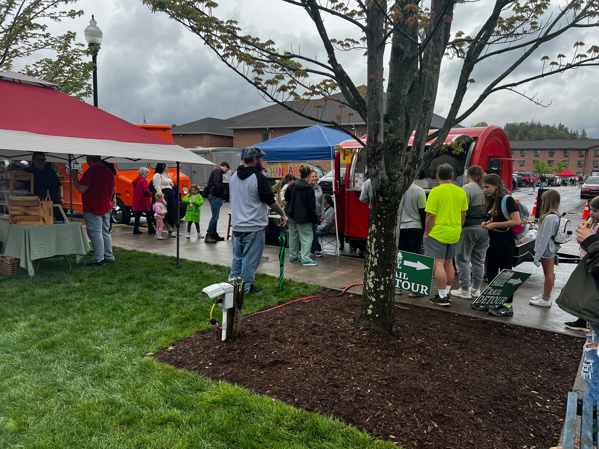 People gather at a market event with food trucks and tents on a cloudy day. Some stand in line, others stroll, and a man with a camera films near a tree in the foreground. The grass and sidewalks are wet.