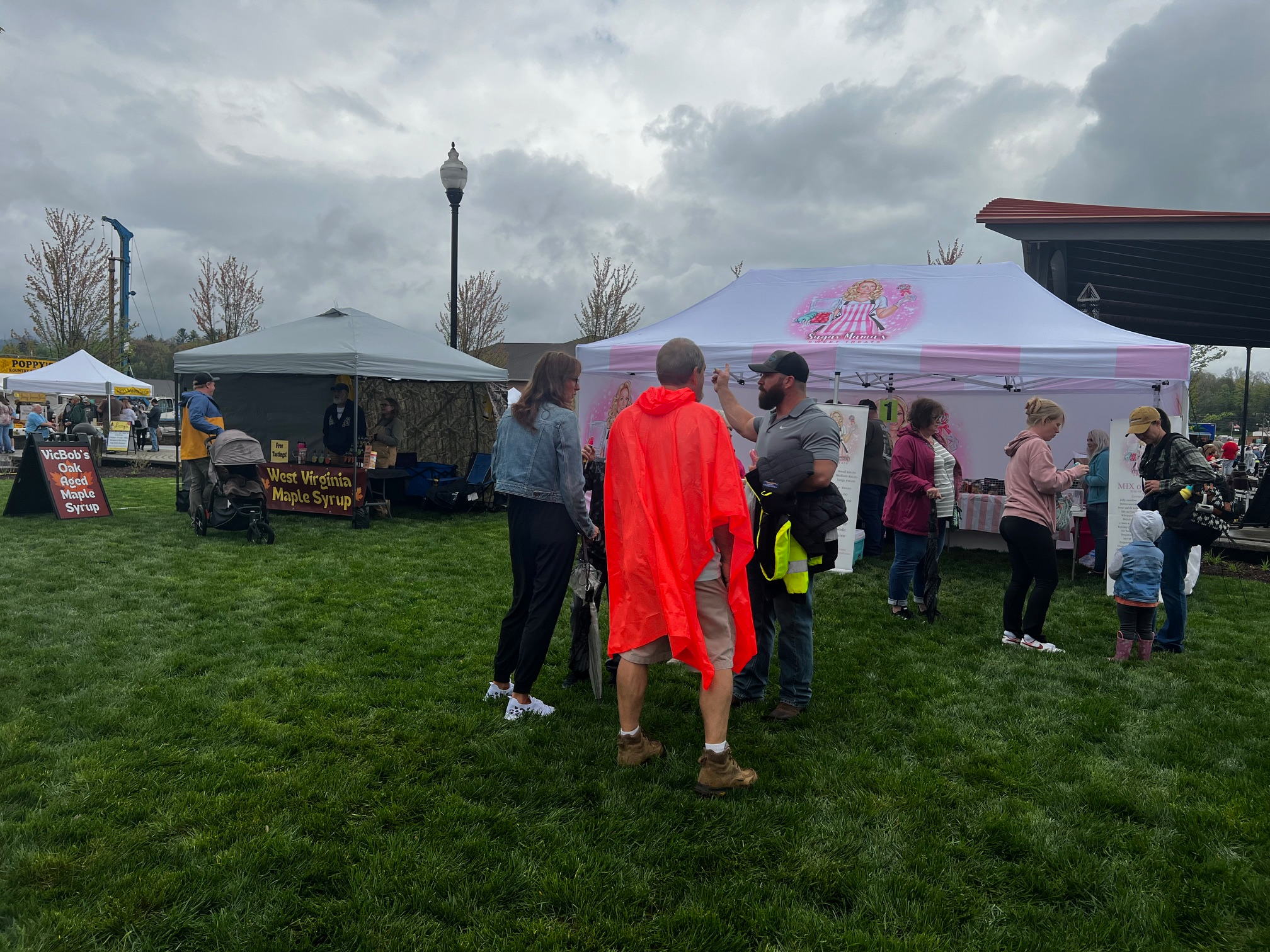 People gather at an outdoor market on a cloudy day, with tents selling West Virginia maple syrup visible. Some are talking, including a person in a bright red rain poncho. The grass is green and trees are budding in the background.