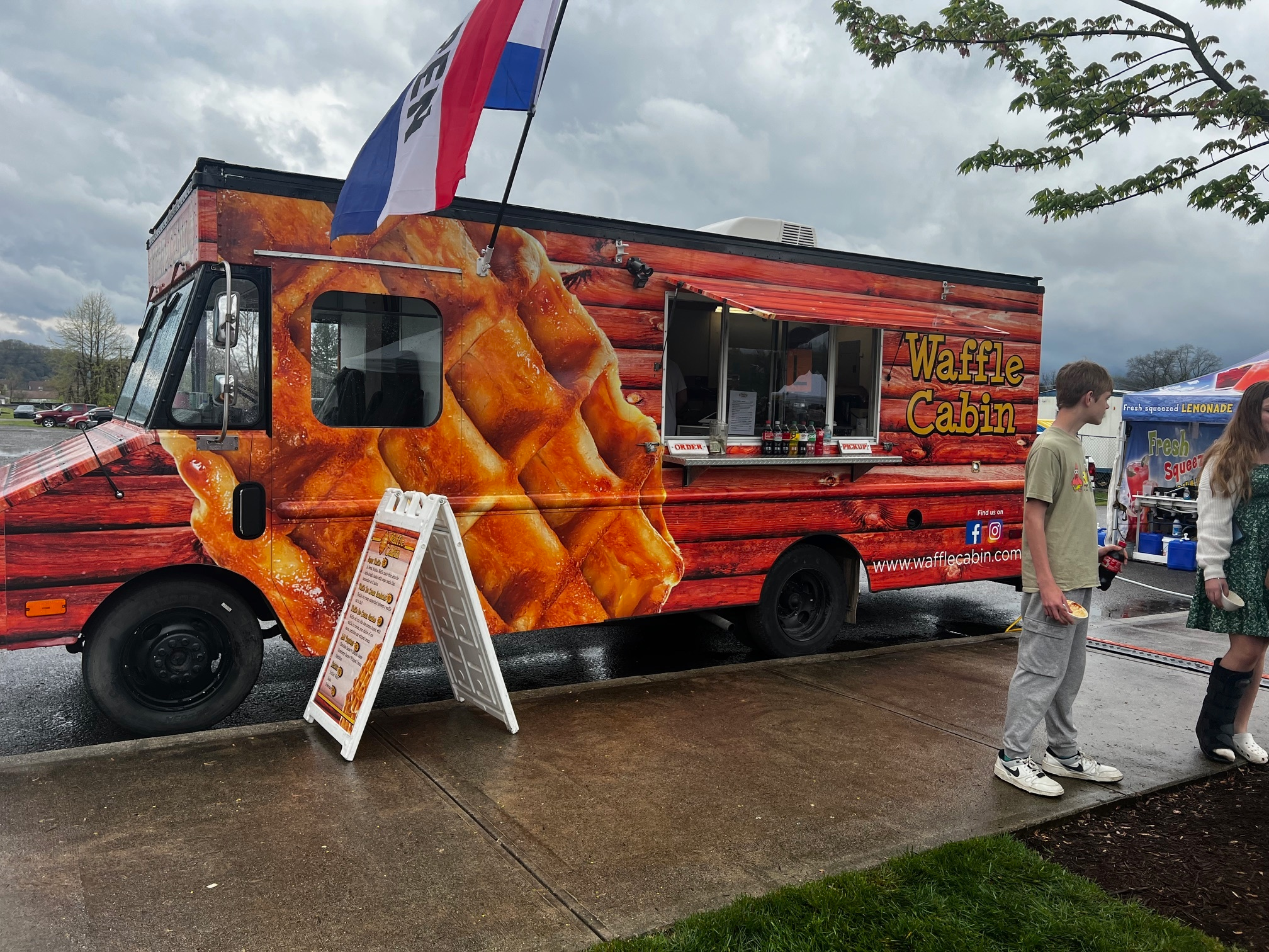 A food truck labeled "Waffle Cabin" with a large waffle image on its side is parked at a curb. Two people stand nearby, and the sky is overcast. A menu board is displayed in front of the truck.