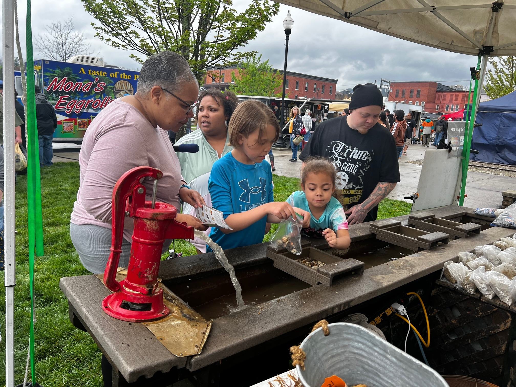 An adult helps three children use a water pump to wash rocks or fossils at an outdoor market or fair. Tents, people, and a "Mona's Eggrolls" food stand are visible in the background.