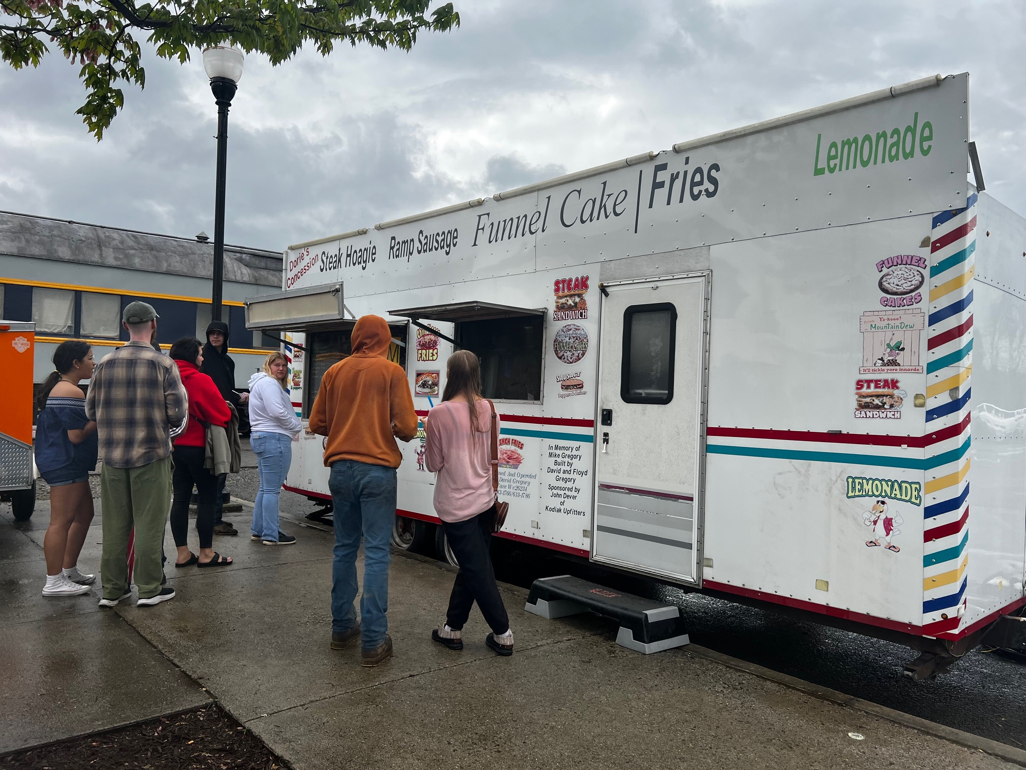 People wait in line at a white food truck selling steak hoagies, sausage, funnel cakes, fries, and lemonade on a cloudy day. The sidewalk is wet, and some people wear hoodies or jackets.