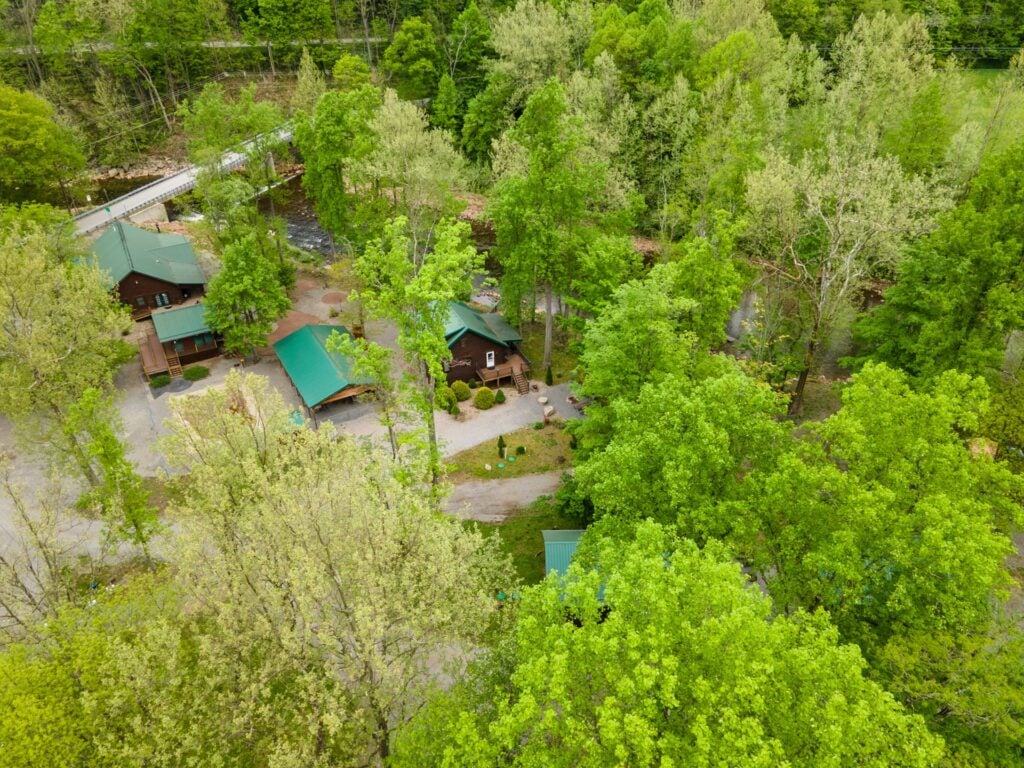 Aerial view of several cabins with green roofs surrounded by dense green trees and intersecting paths in a wooded area.