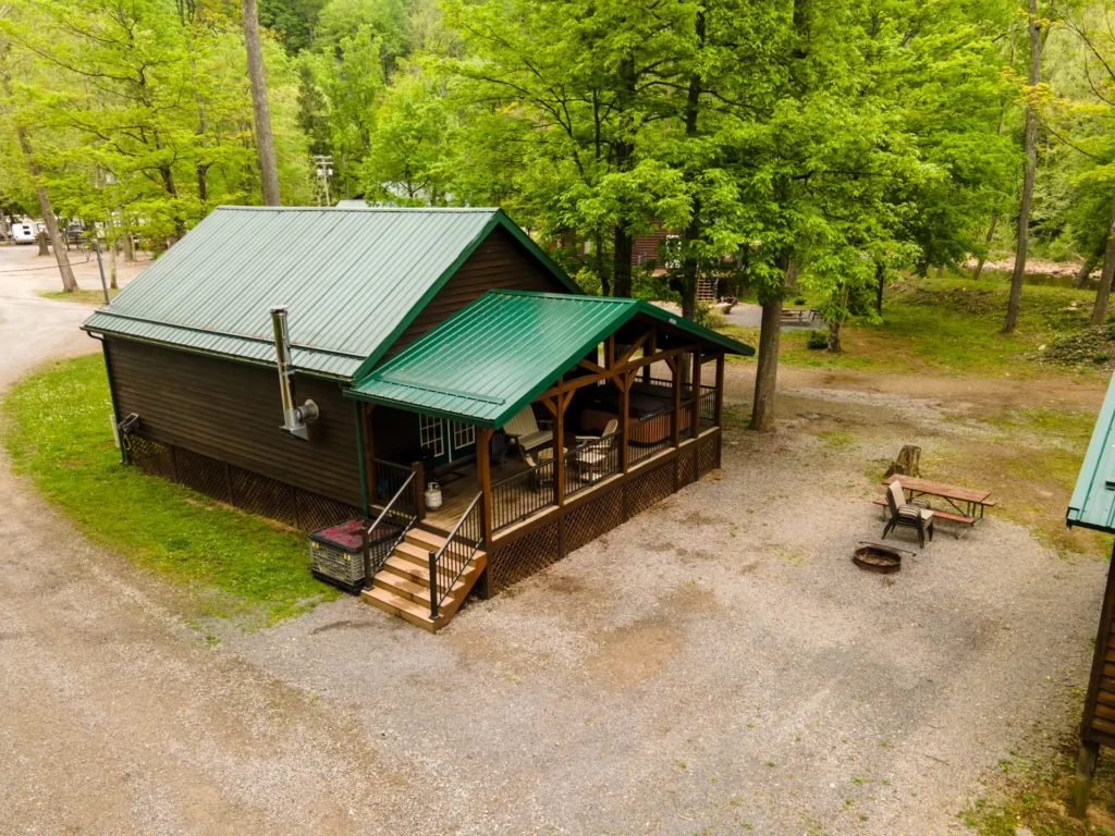 A wooden cabin with a green metal roof sits among trees, featuring a covered porch, picnic table, and fire pit on a gravel lot.