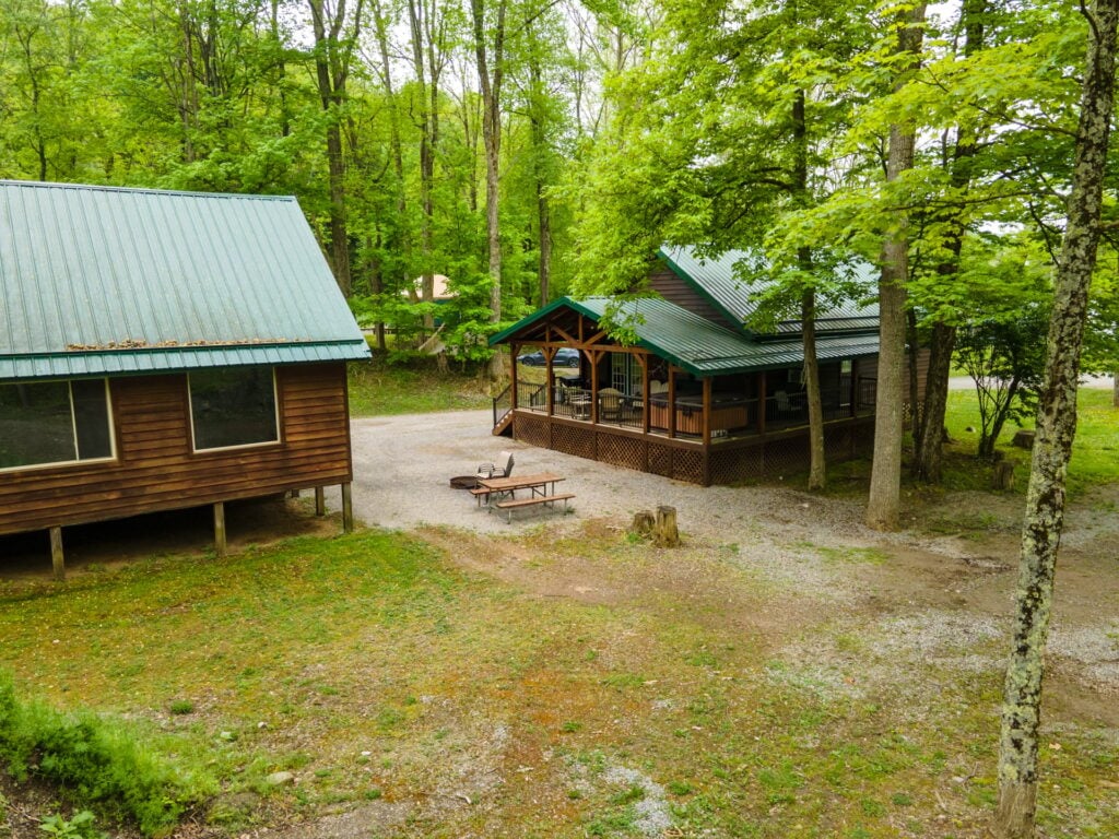Two wooden cabins with green metal roofs sit among trees in a wooded area, with a gravel ground, picnic table, and a fire pit between them.