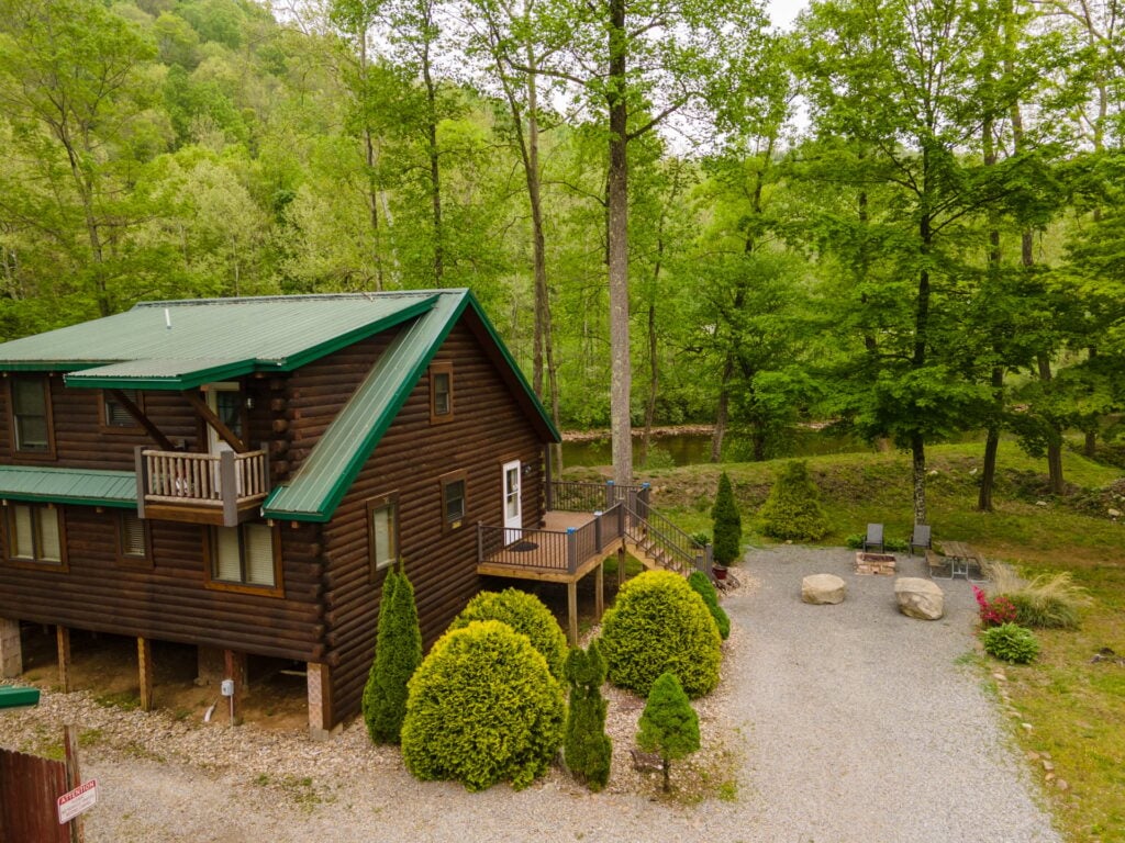 A log cabin with a green roof is surrounded by trees, gravel pathways, shrubs, and outdoor seating near a small river in a wooded area.