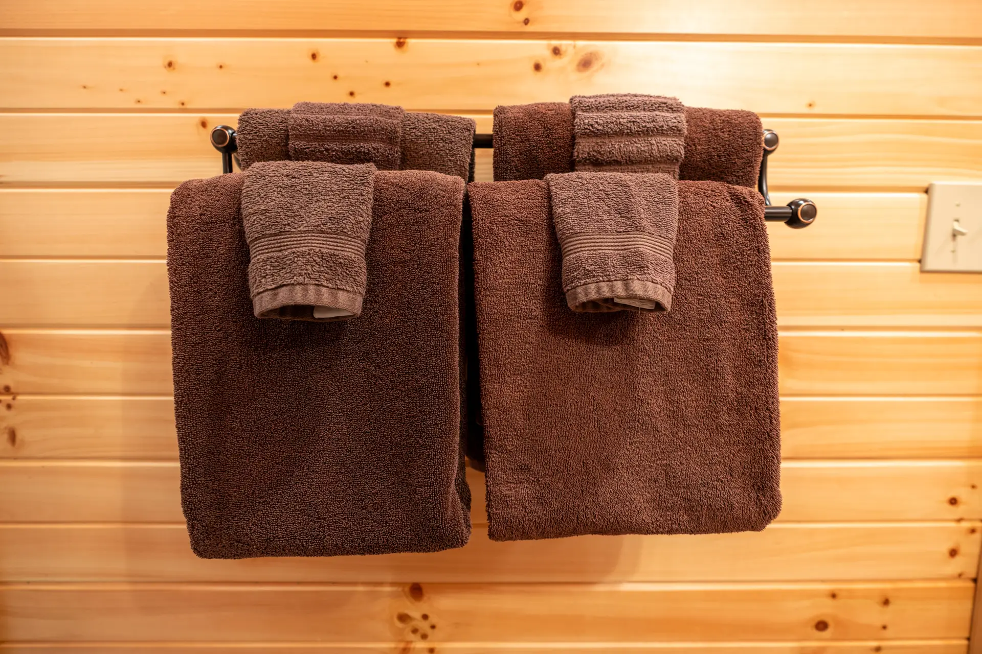 Two brown towels and two matching hand towels neatly folded and hanging on a black towel rack against a wooden wall.