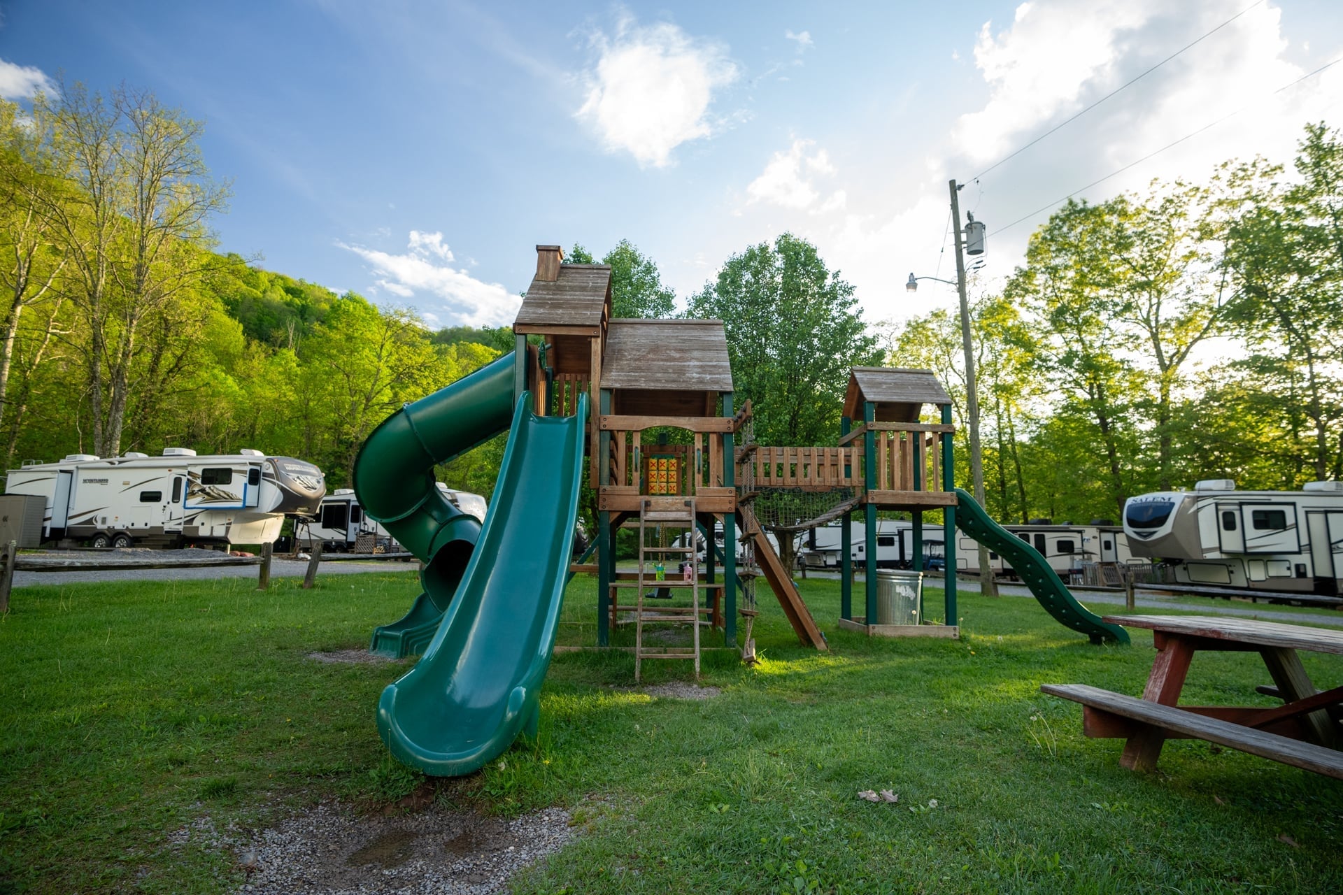 Wooden playground with green slides and climbing features, surrounded by grass, picnic table, trees, and several RVs in the background.
