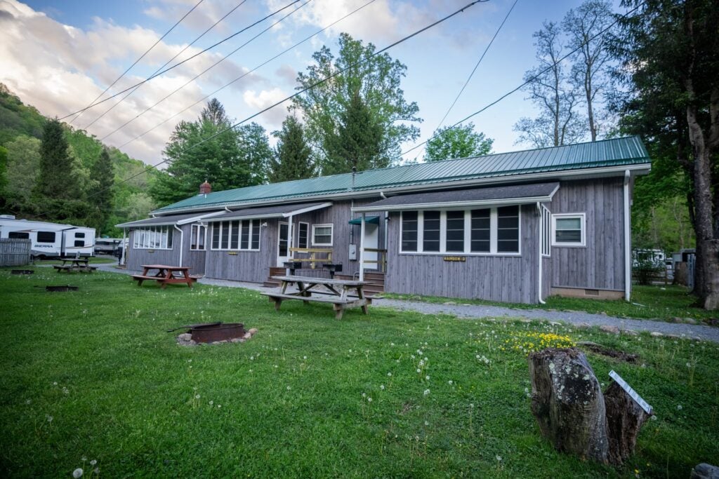 A single-story wooden cabin with a green metal roof, surrounded by grass, picnic tables, and trees, with an RV visible in the background.