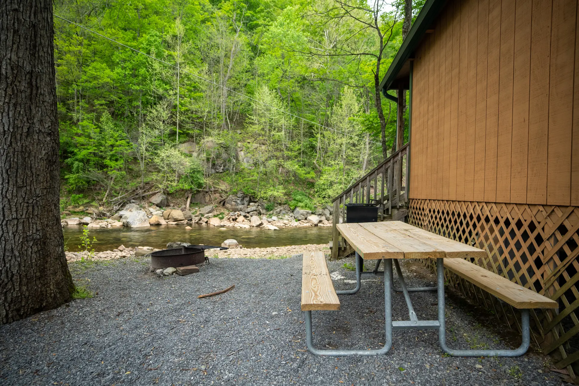 Wooden picnic table and fire pit on gravel beside a cabin, overlooking a rocky stream and surrounded by green trees.