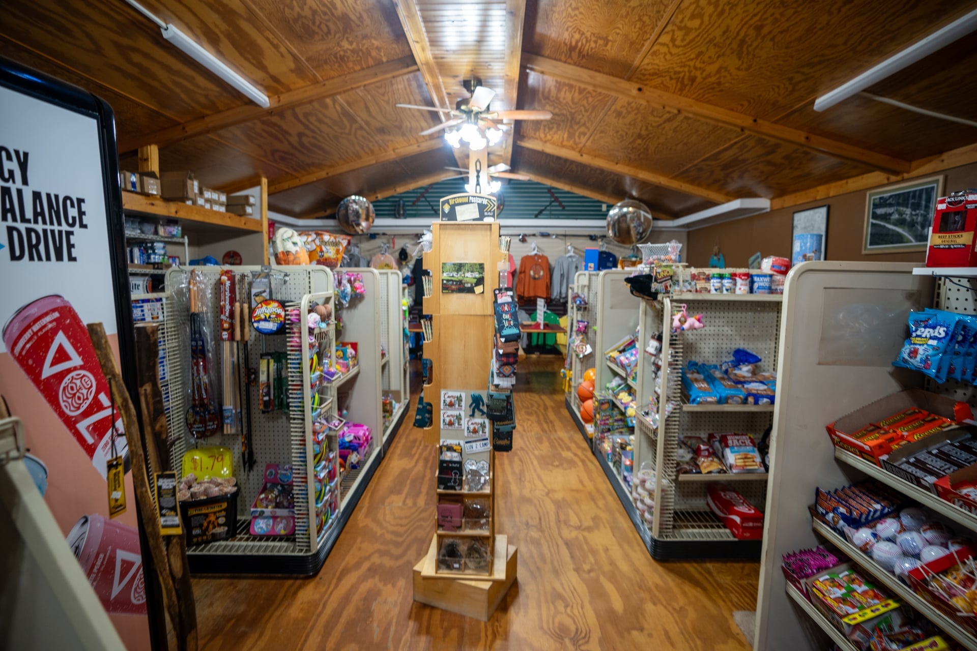 A small convenience store interior with wooden floors, shelves stocked with snacks and assorted items, and a rack of souvenirs in the center aisle.