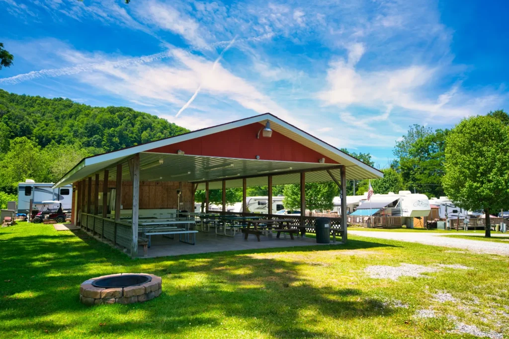 Open-air pavilion with picnic tables at a campground, surrounded by green grass, trees, RVs, and a fire pit under a blue sky with scattered clouds.