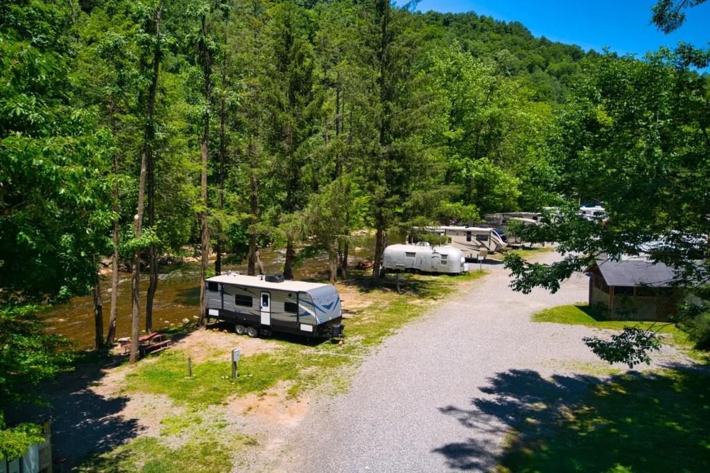 A gravel road runs through a tree-filled campground with several parked RVs and trailers, bordered by a river and surrounded by greenery.