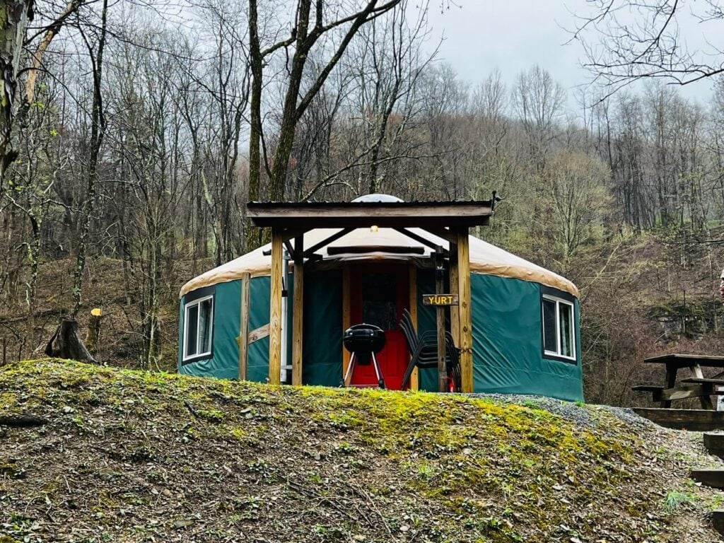 A green and tan yurt with a red door sits in a wooded area; a grill and chairs are under a small wooden shelter at the entrance. Trees without leaves surround the yurt.