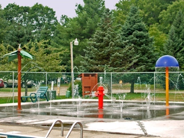 Outdoor splash pad with water jets, a red hydrant, and colorful spray features, surrounded by a chain-link fence and trees in the background.