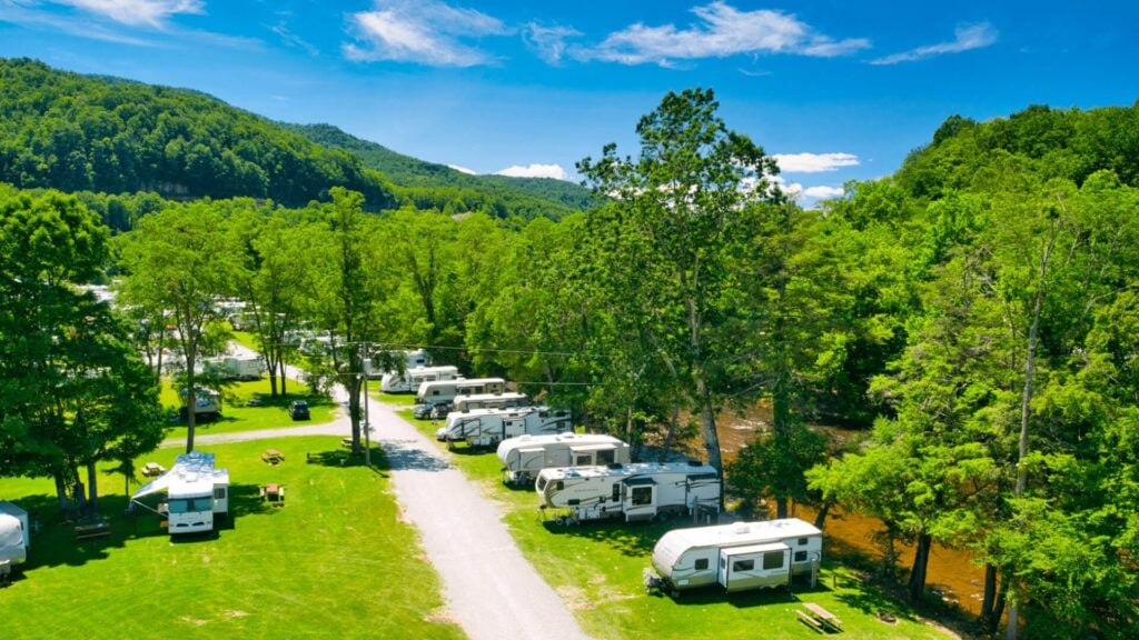 Rows of RVs parked along a gravel road in a green, tree-filled campground near a river with mountains in the background under a blue sky.