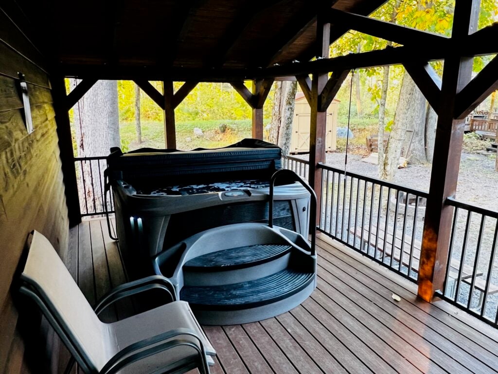 A covered wooden deck with a hot tub, two steps leading up to it, and a white bench; surrounded by trees and a fenced yard.