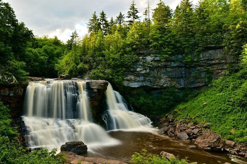 A wide waterfall cascades over a rocky ledge into a river, surrounded by dense green trees and a steep stone cliff.
